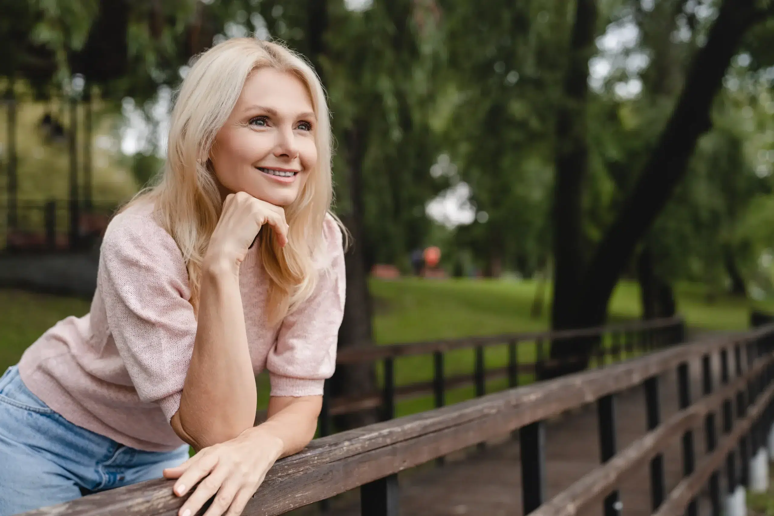 Woman smiling outdoors, leaning on a wooden railing in a park setting.